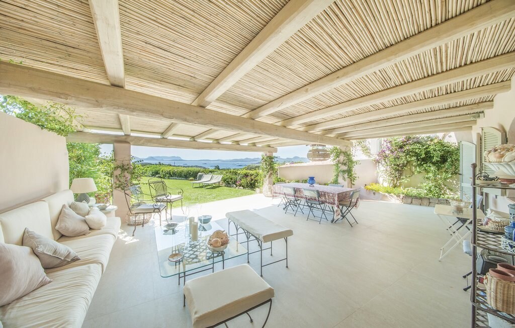 A spacious outdoor patio is seen, featuring a wooden ceiling with exposed beams. Comfortable seating arrangements include a sectional sofa and a dining table set. Lush greenery can be noted in the background, along with views of rolling hills and a distant sea.