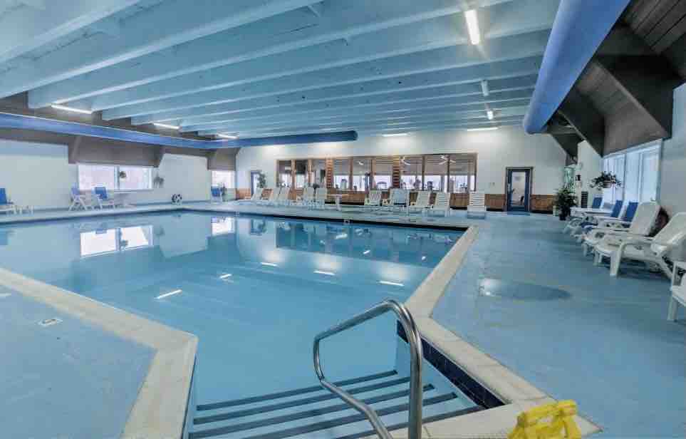 An indoor pool area is visible, featuring clear blue water and a spacious deck surrounded by lounge chairs. Large windows allow natural light to fill the space, highlighting the high ceiling and bright blue accents on the walls.
