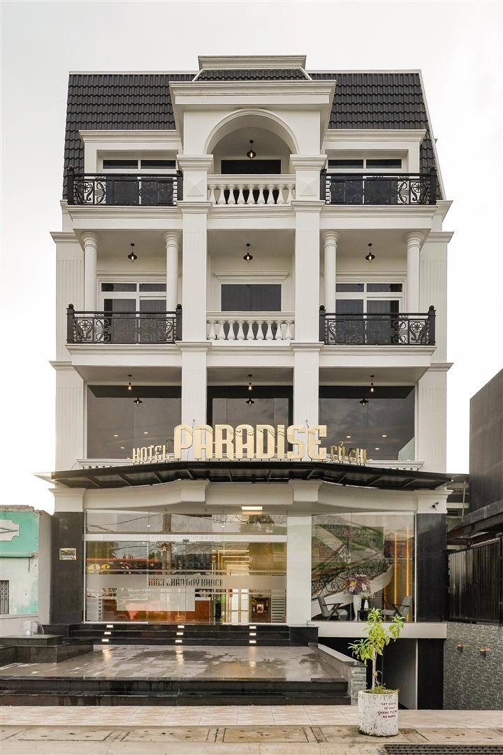 The exterior of Paradise Hotel showcases a modern design with three stories, featuring a combination of large glass windows and decorative balconies. A glass entrance allows visibility into the hotel's inviting lobby area. The structure is accented with minimalist landscaping beside the entrance.