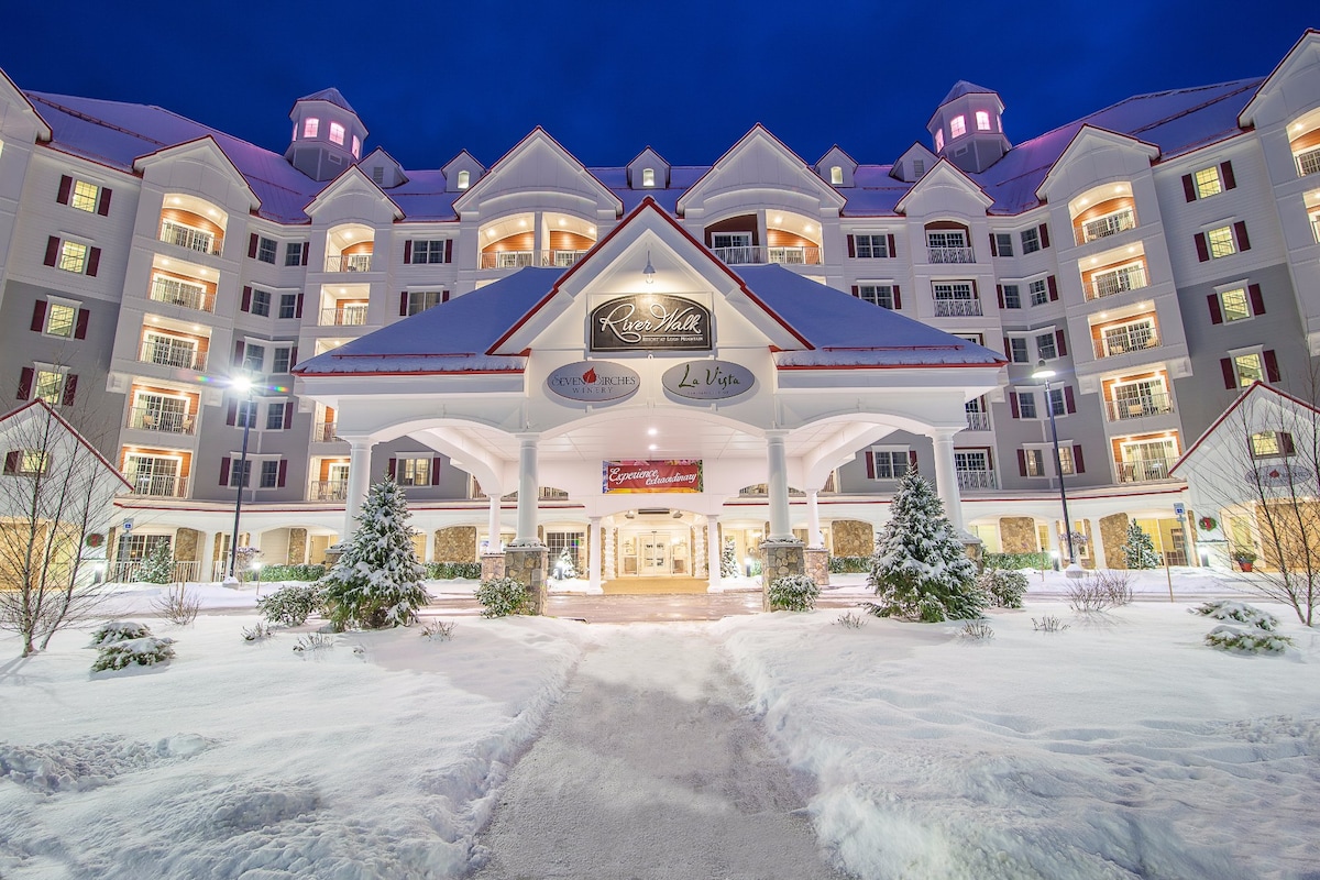 A grand entrance to Riverwalk Resort is showcased, framed by freshly fallen snow. The building features multiple stories with a peaked roof, large windows, and decorative architectural elements. Snow-covered shrubbery and a well-maintained walkway lead to the welcoming entrance underneath a large canopy.