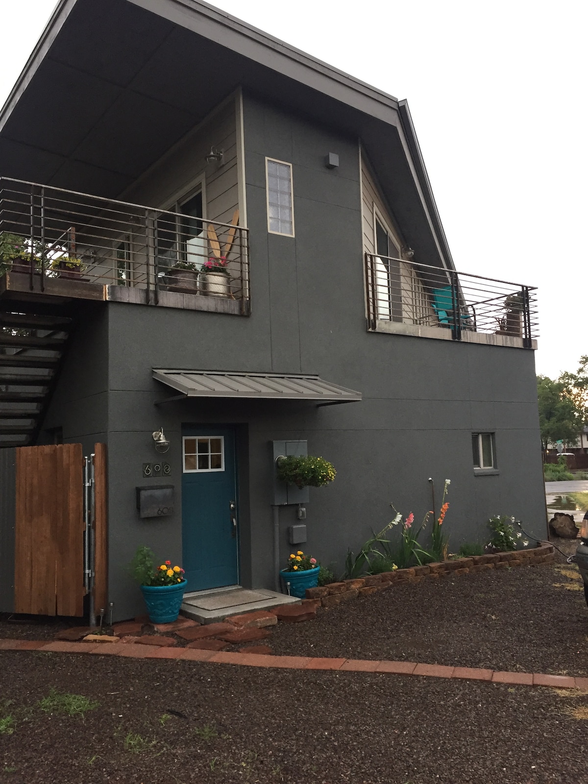 A modern two-story home features a distinctive angular design and a combination of grey exterior walls and a metal roof. The entryway is highlighted by a vibrant blue door, flanked by flower pots. A staircase leads to an upper level balcony adorned with greenery.