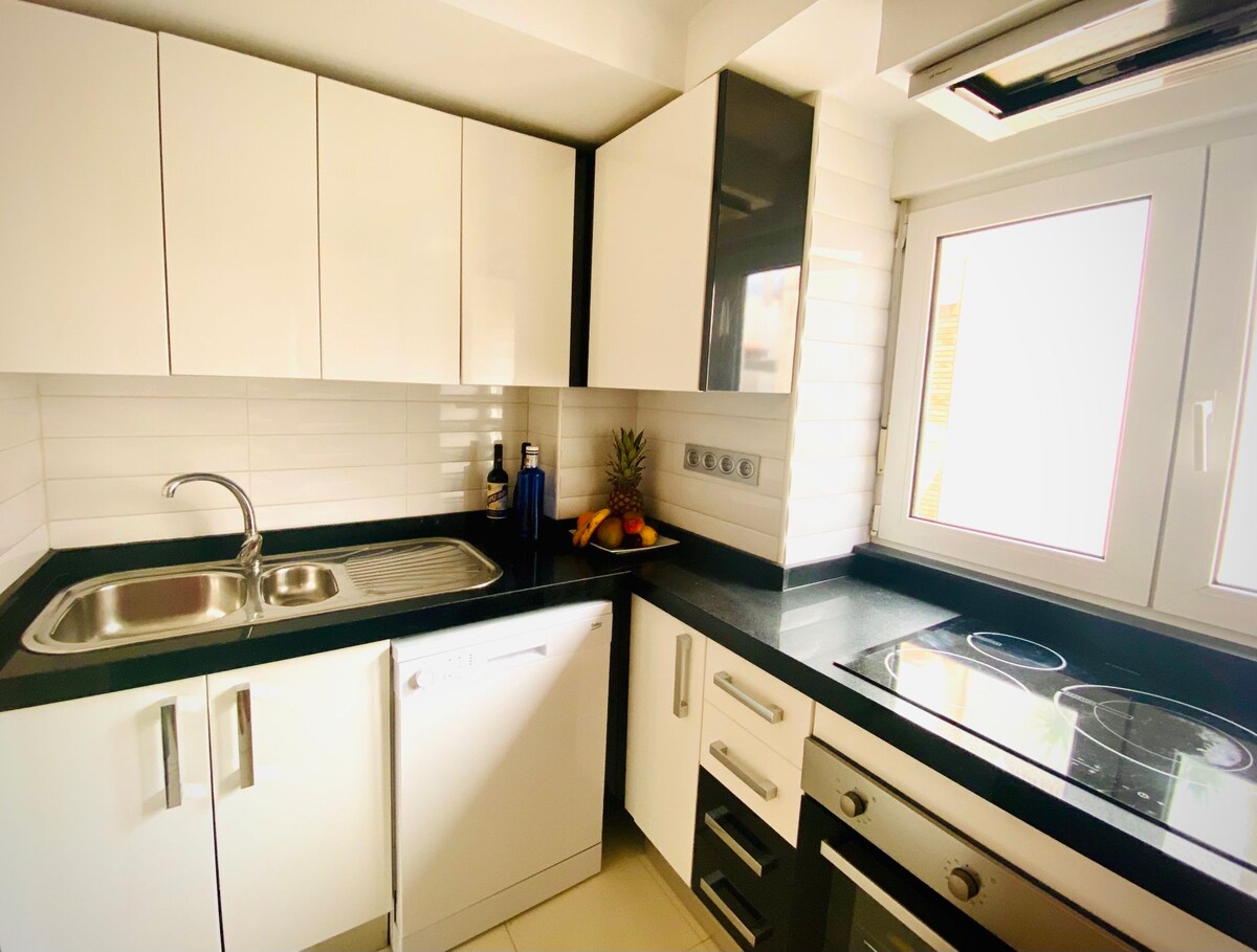 A modern kitchen features sleek black granite countertops complemented by white cabinetry. A stainless steel sink and dishwasher are visible, alongside a built-in stove and oven. Natural light enters through a window, illuminating a small fruit display on the counter.