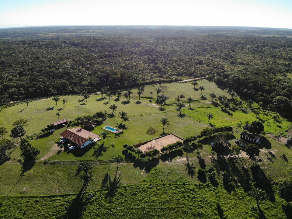 An aerial view of a spacious property reveals a well-maintained green landscape featuring palm trees. A swimming pool is visible alongside a tennis court, with a furnished house positioned centrally, surrounded by open fields and natural scenery.
