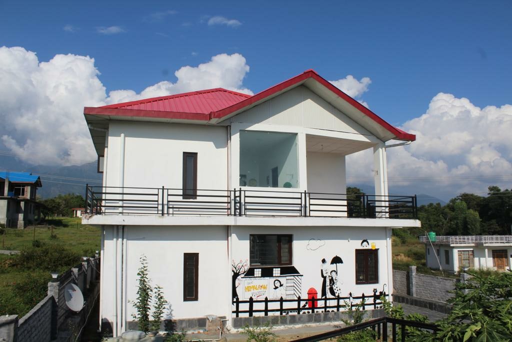 A modern two-story mansion is displayed, featuring a striking red roof and large glass balconies. A mural adds a playful touch to the exterior, while surrounding greenery complements the scenic backdrop of the Himalayas, creating a serene and welcoming atmosphere.