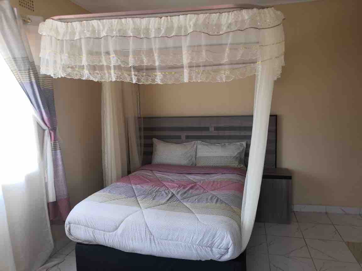 A spacious bed is positioned under a decorative canopy, adding an elegant touch to the sleeping area. Light enters through a nearby window, illuminating the soft colors of the bedding and the neutral tones of the walls.