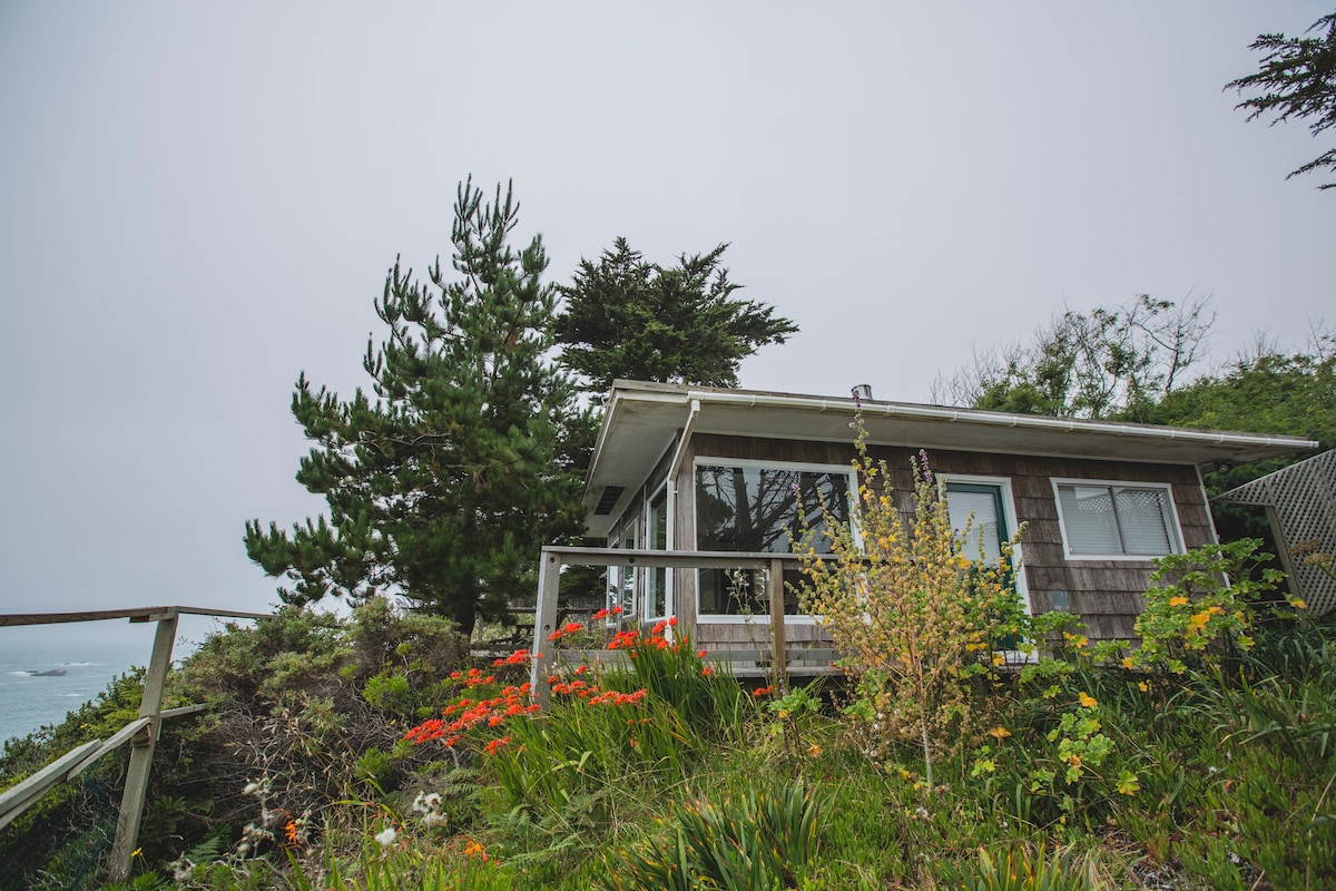 A cottage is situated on a slight incline, surrounded by greenery and colorful flowers. Large windows offer views of the ocean, while trees provide a natural backdrop against a cloudy sky.