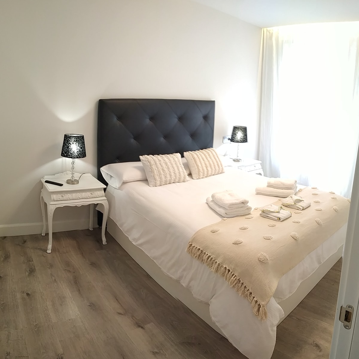 A spacious sleeping area is presented with a neatly made bed featuring soft white linens and a decorative beige throw. Flanking the bed are two elegant nightstands with black lamps. Natural light filters in through sheer curtains, creating a bright and calming ambiance.