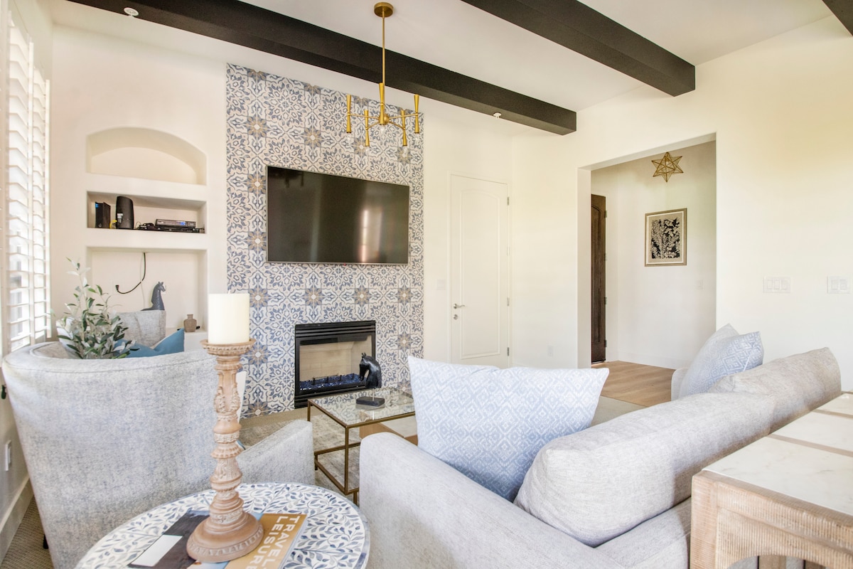 The living area features a large, comfortable sectional sofa, a coffee table with decorative accents, and a modern gas fireplace. A mounted flat-screen TV is visible above the fireplace, with patterned tile accents on the wall. Natural light floods the space through window shutters.