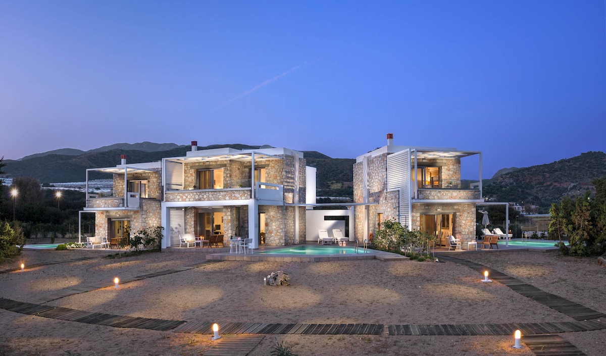 The exterior of Cedar Bay Villas showcases a modern, two-storey stone structure nestled in a sandy landscape. Soft lighting highlights the architectural details at dusk, while pathways lead to each villa. The surrounding area features landscaped greenery and a swimming pool visible in the center.
