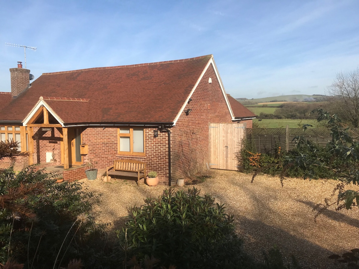 A charming bungalow is situated amidst lush greenery, showcasing a gravel driveway leading to a wooden front door. A wooden bench is placed outside, inviting relaxation, while gentle hills are visible in the background, enhancing the natural setting.