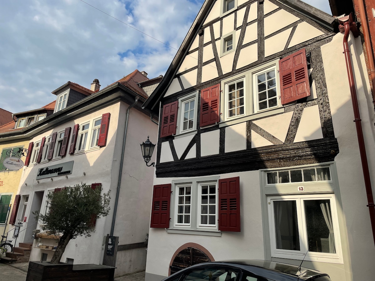 A historic building is shown with black and white timber framing, accented by red shutters on the windows. The structure is set in a quaint street, featuring a nearby lamp post and a glimpse of a neighboring building with decorative elements.
