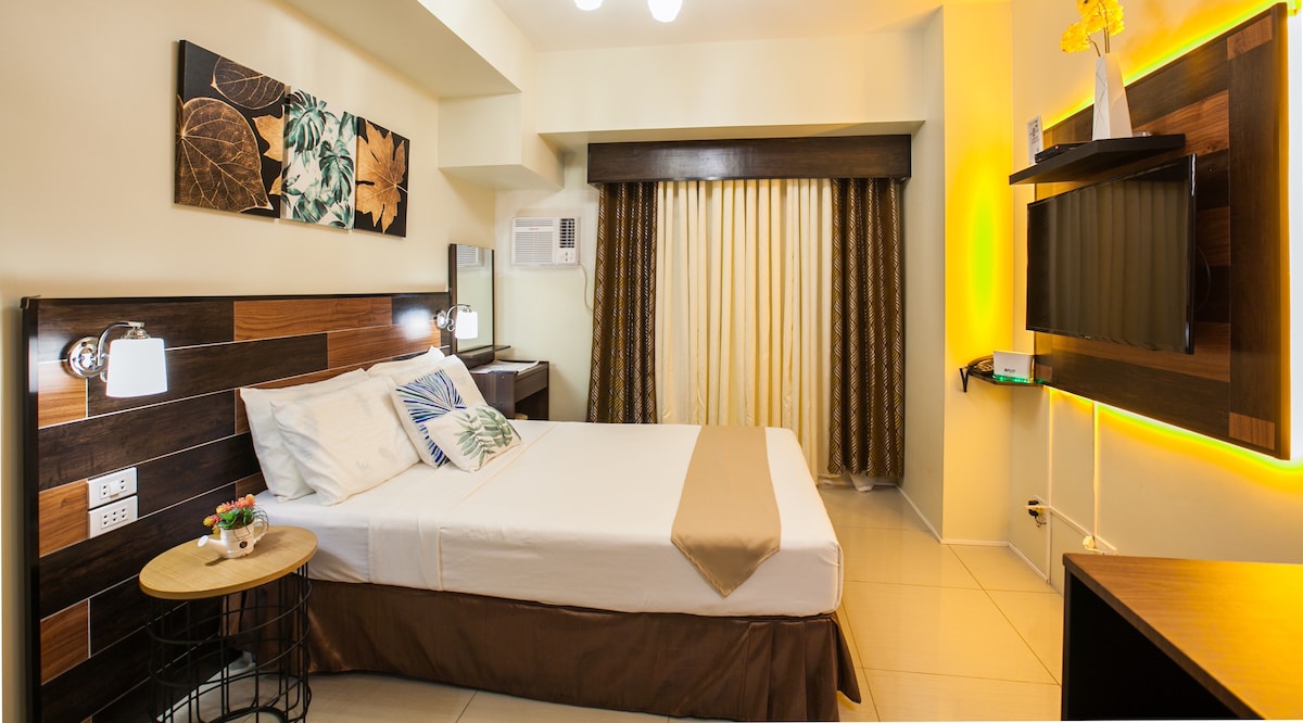 The studio unit features a double bed with decorative pillows against a wood-paneled headboard. Natural light filters through sheer drapes, while a compact side table adds functionality. A wall-mounted television is positioned across from the bed, providing a modern touch to the space.