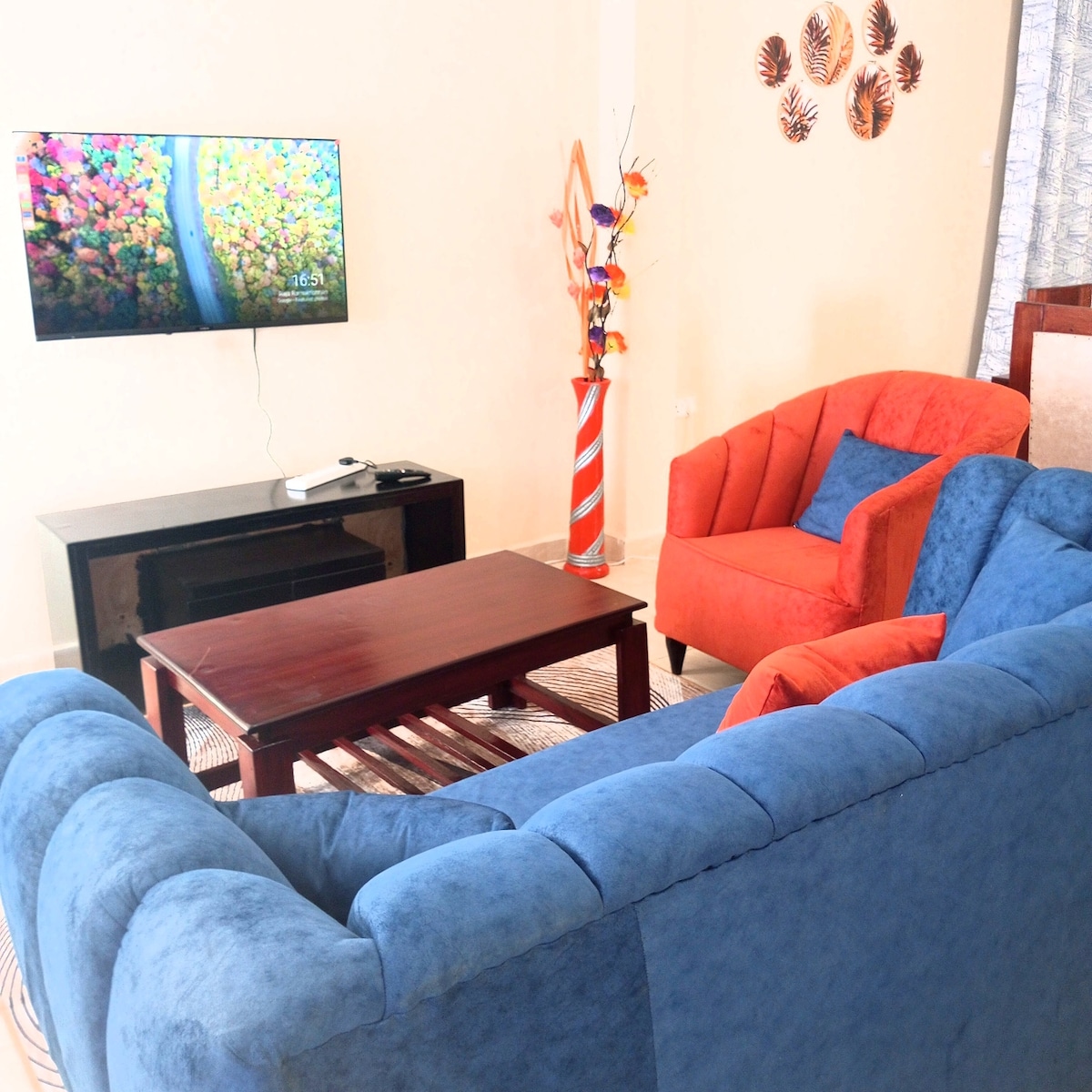 A cozy living area features a blue sofa and an orange armchair arranged around a wooden coffee table. A modern television is mounted on the wall, displaying a vibrant nature scene. Decorative elements, such as a floor vase with flowers, add a touch of color.