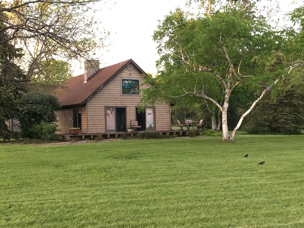 The exterior of the A-frame cottage is shown, featuring a rustic wood-shingled façade with a sloped roof. A large lawn surrounds the house, complemented by a mature tree. The warm light of the evening sun adds a serene ambiance to the setting.