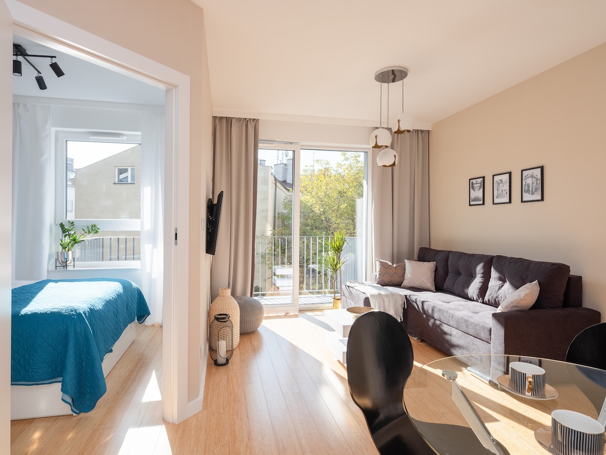 The living area features a comfortable dark sofa and a round dining table set for four, with sunlight streaming through large sliding doors. A cozy blue blanket adorns the nearby sleeping area, which is partially visible. Plants softly complement the natural light-filled space.