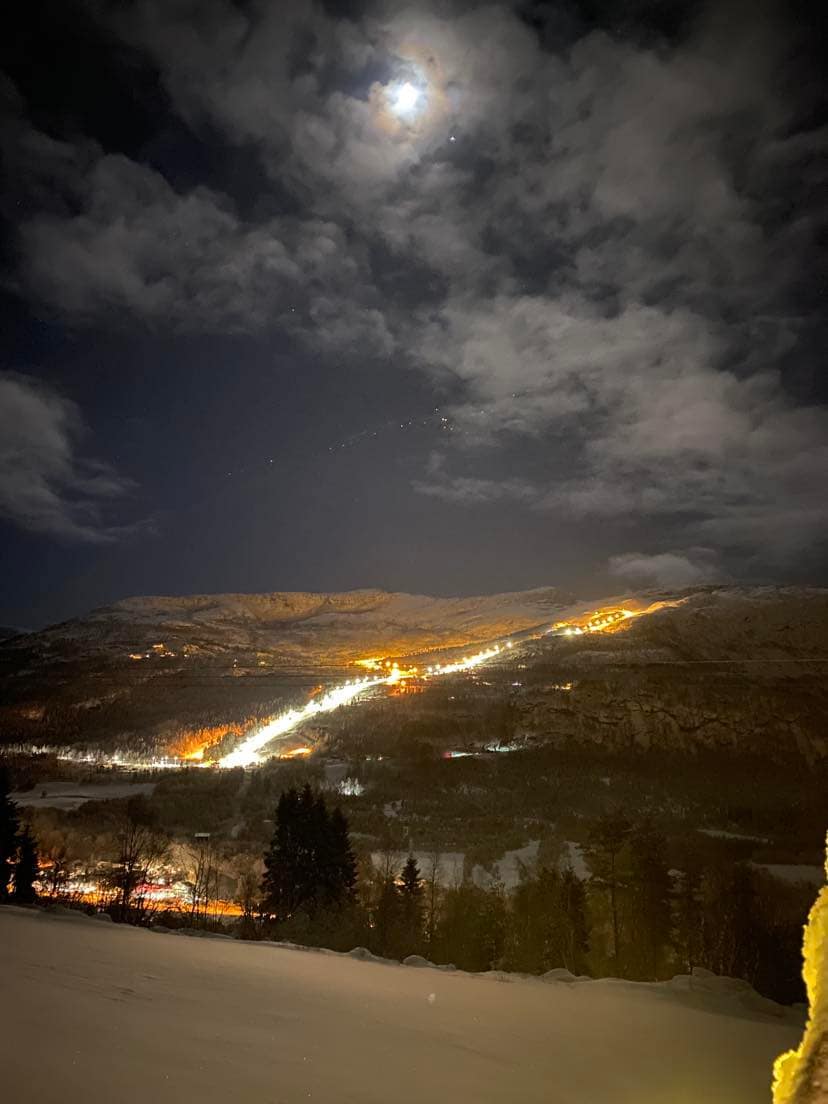 A night scene showcases a mountainous landscape illuminated by soft lights from a ski slope. The moon casts a gentle glow over the clouds, while the snowy terrain is visible in the foreground. Pine trees frame the view, enhancing the serene atmosphere.
