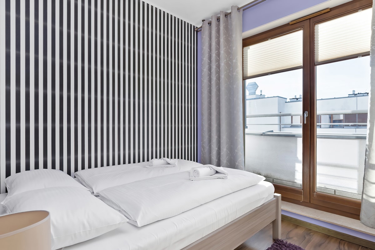 Spálňa s veľkým oknom a tapetou s čiernobielymi pruhmi. V popredí je posteľ a interiér je moderný a minimalistický.