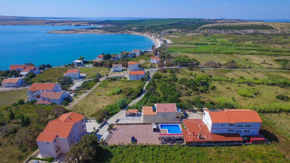 An aerial view showcases a tranquil landscape featuring the coastline, lush greenery, and well-spaced buildings with red rooftops. A large swimming pool is visible surrounded by a terrace, and the serene blue waters contrast with the surrounding land.