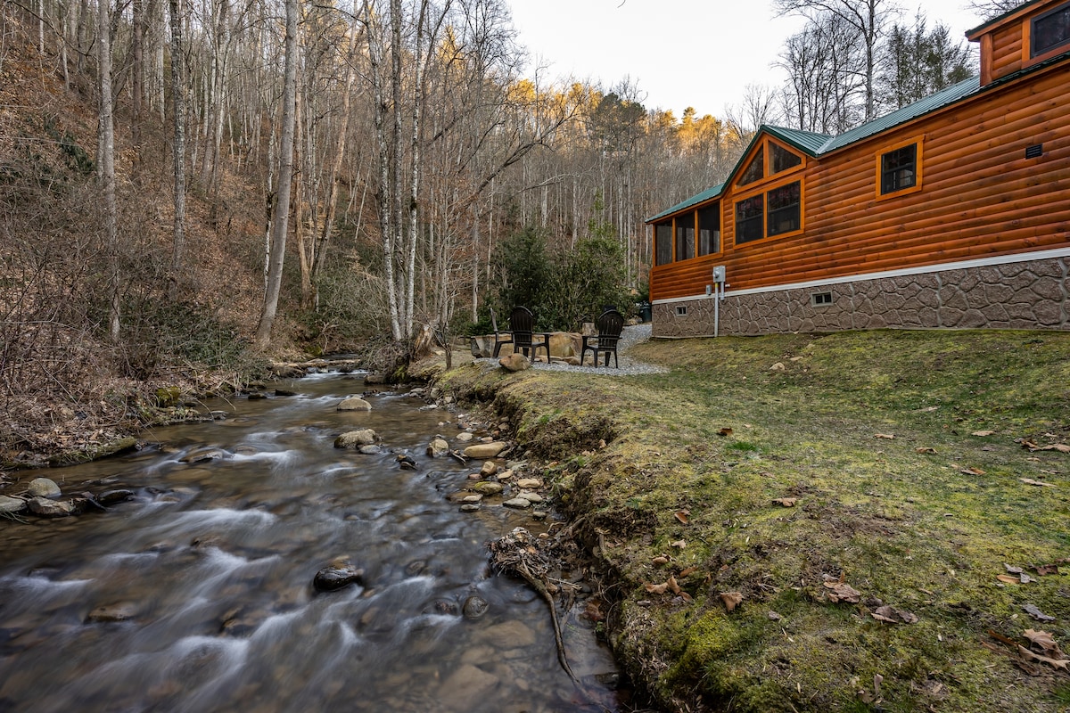 The exterior of a tiny home is seen alongside a softly flowing creek, bordered by a grassy area. A firepit and seating arrangement are present, surrounded by trees, adding to the natural setting. The wooden structure features large windows that look out onto the serene landscape.