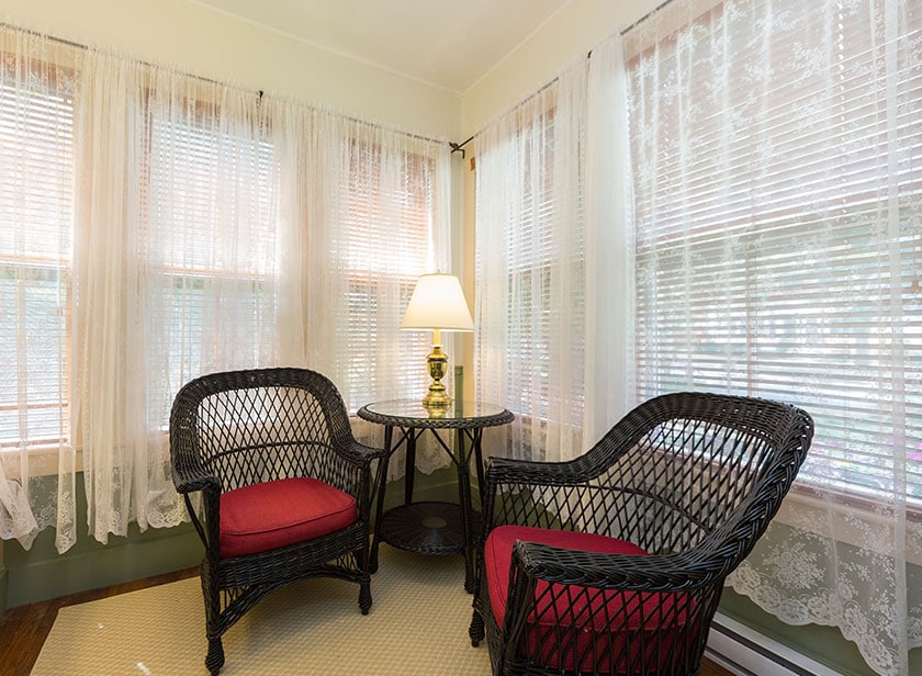 A cozy sitting area features two wicker chairs with red cushions positioned around a small table. Soft light is provided by a lamp on the table. Sheer white curtains cover the windows, allowing diffused natural light to enter the room.