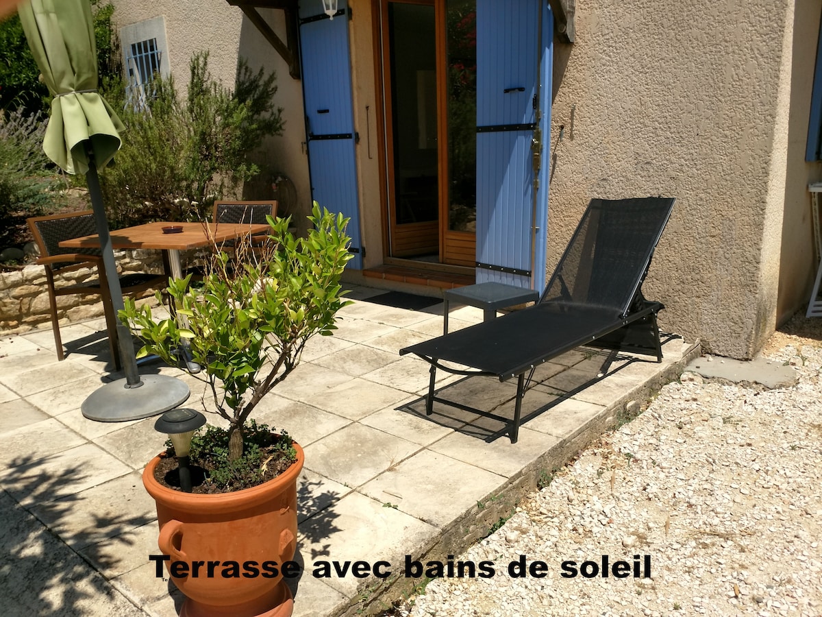 A small terrace area is featured, furnished with a dining table and chairs. A sun lounger is positioned nearby, alongside a potted plant. The exterior doorway showcases blue shutters, leading into the apartment.
