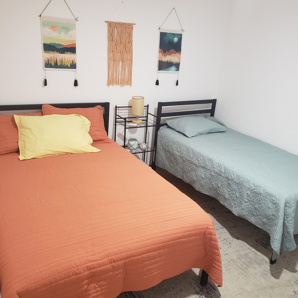 The room features two beds, one with an orange coverlet and the other with a light blue quilt. Colorful wall hangings are displayed above the beds, and a small shelf is positioned between them, showcasing decorative items.
