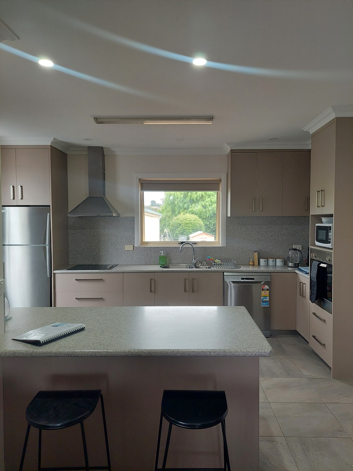 A modern kitchen features sleek cabinetry with light brown finishes and a spacious countertop. Stainless steel appliances, including a refrigerator and a microwave, are included. A window offers natural light and a view of greenery outside, enhancing the kitchen's functionality.