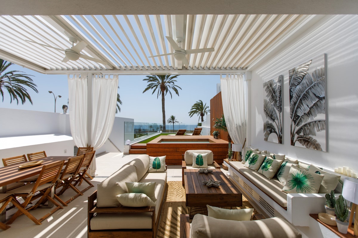 An outdoor living area features a large dining table and a comfortable seating arrangement. Ceiling fans circulate air among the modern furnishings. Sheer curtains frame the space, while palm trees and sea views are visible in the background, offering a serene ambiance.