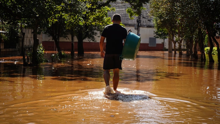 Uma pessoa com cabelos escuros bem curtos se afasta, carregando uma banheira de plástico por uma rua inundada.