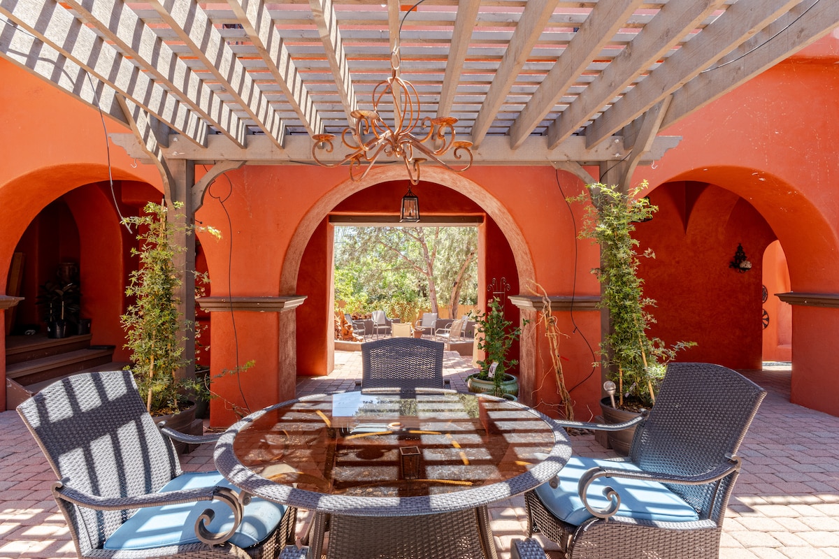 An outdoor dining area features a round table surrounded by four chairs, located under a wooden pergola. The warm orange walls and lush greenery create a serene backdrop, while additional seating is visible in the area beyond, enhancing the inviting atmosphere.