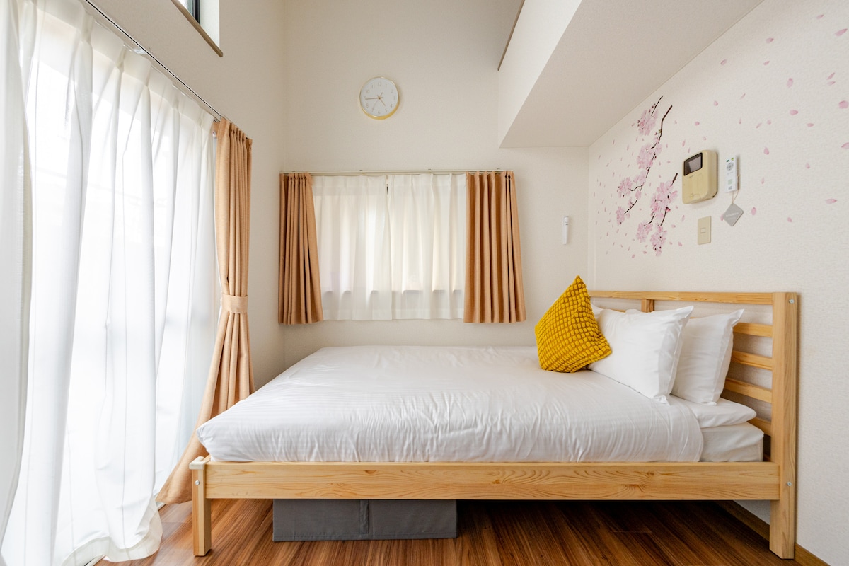 A cozy sleeping area features a spacious bed with crisp white linens. A vibrant yellow cushion adds a pop of color. Natural light streams through large windows adorned with flowing curtains, creating a warm and serene environment.