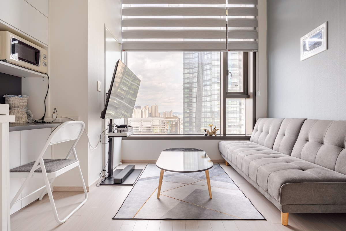 A bright and airy living space features a comfortable gray sofa with a sleek coffee table set on a modern area rug. Large windows provide an expansive view of the city skyline, while a television and a compact kitchenette are conveniently positioned nearby.