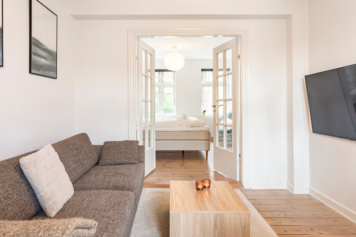 A living area is presented with a contemporary gray sofa positioned across from a wooden coffee table. Soft lighting highlights the entrance to a spacious bedroom, where a large bed is visible through the open double doors. Textured art pieces adorn the walls.