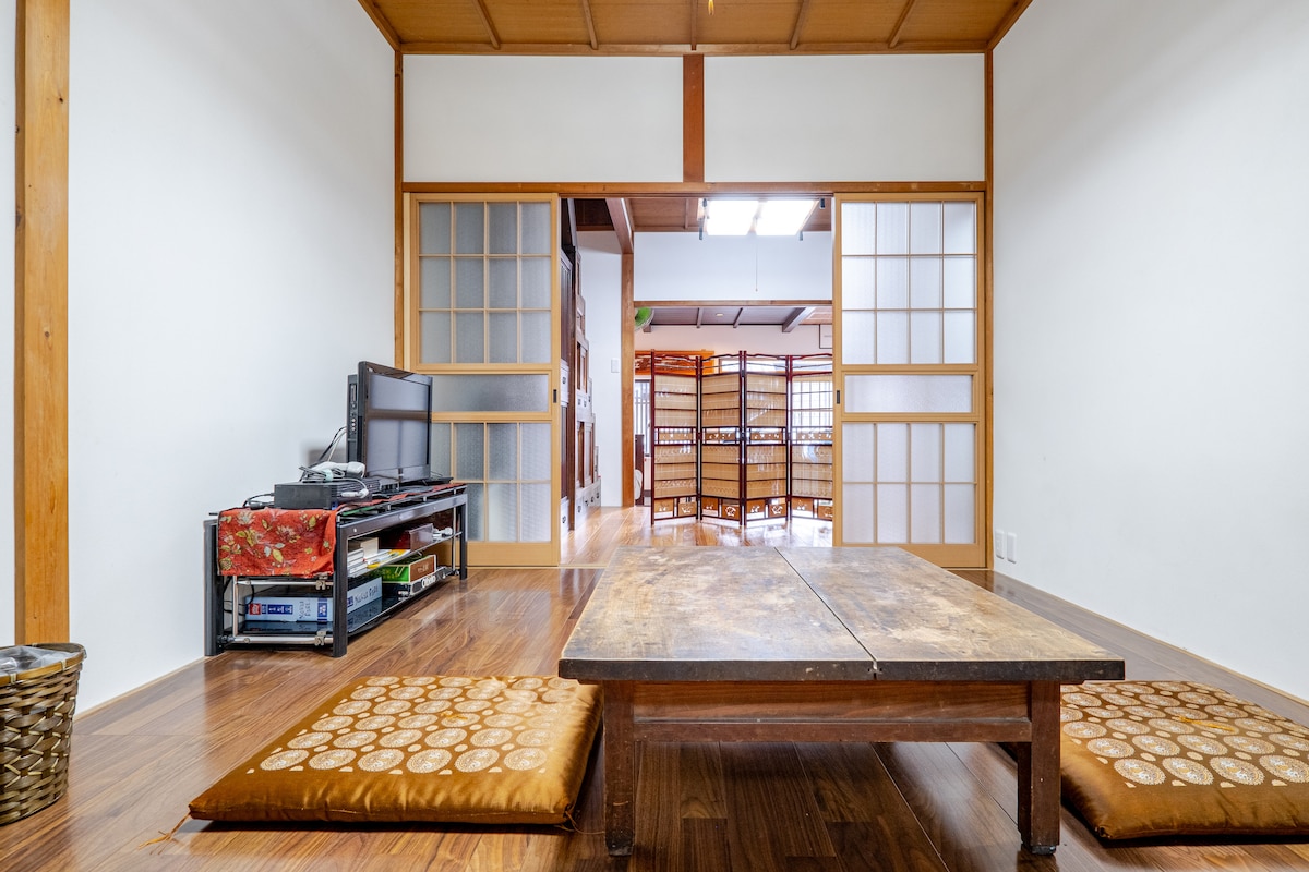 A traditional Japanese room features a low wooden table surrounded by floor cushions. A television stand is positioned nearby, and sliding shoji doors lead to an adjacent space. The warm wood flooring complements the natural wood accents of the room.