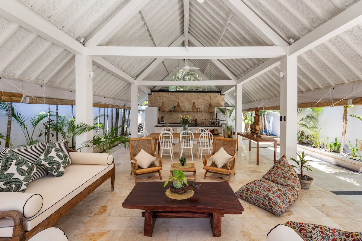 An open and airy lounge area is illuminated by natural light, featuring a high ceiling with wooden beams. Comfortable seating includes a spacious sofa and several chairs, arranged around a wooden coffee table. Tropical plants are placed throughout, enhancing the serene ambiance.