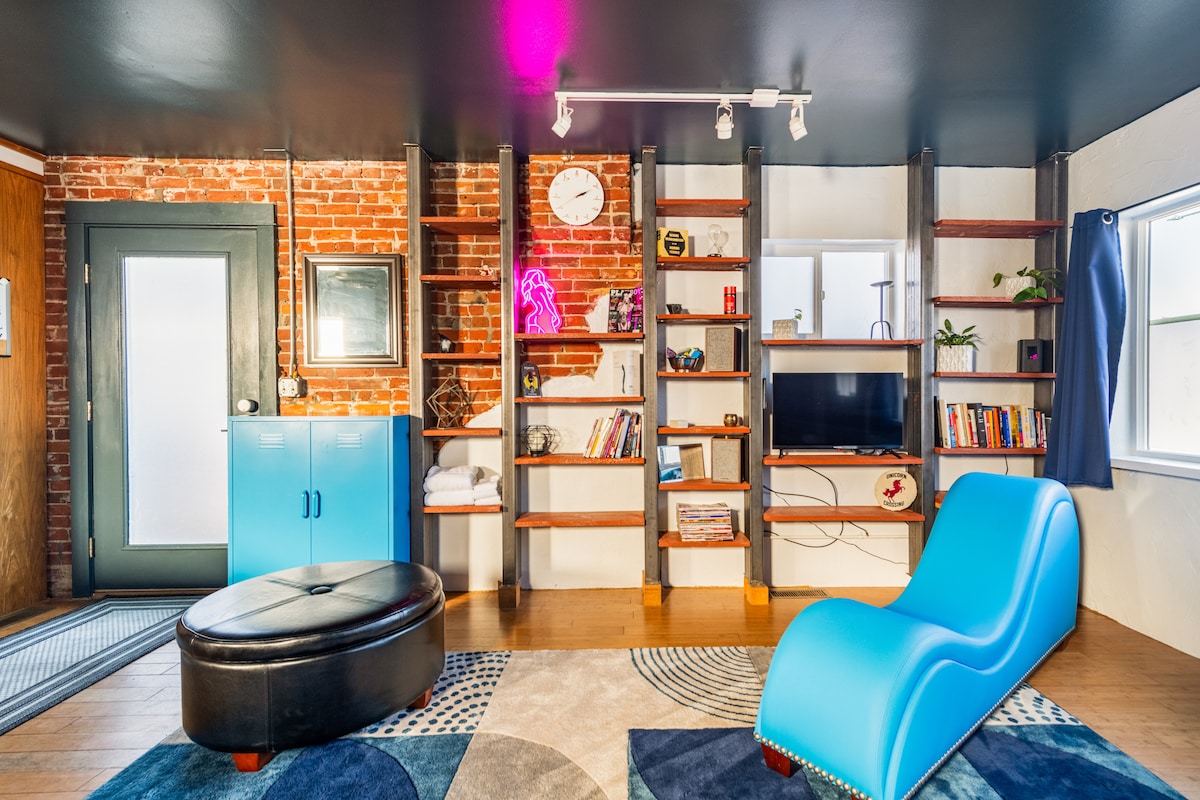 A cozy living area features a vibrant blue Tantra Chair alongside a black ottoman. A striking brick wall contrasts with the sleek black ceiling. Shelves display books and decorative items, while a clock and neon accent light add character. A television is positioned near a window.