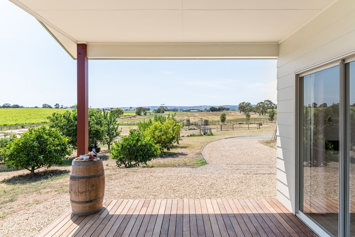 An outdoor deck area is featured, bordered by wooden decking and a barrel table. A spacious view showcases vibrant green vineyards and open countryside, with trees and shrubs dotting the landscape under a clear sky.