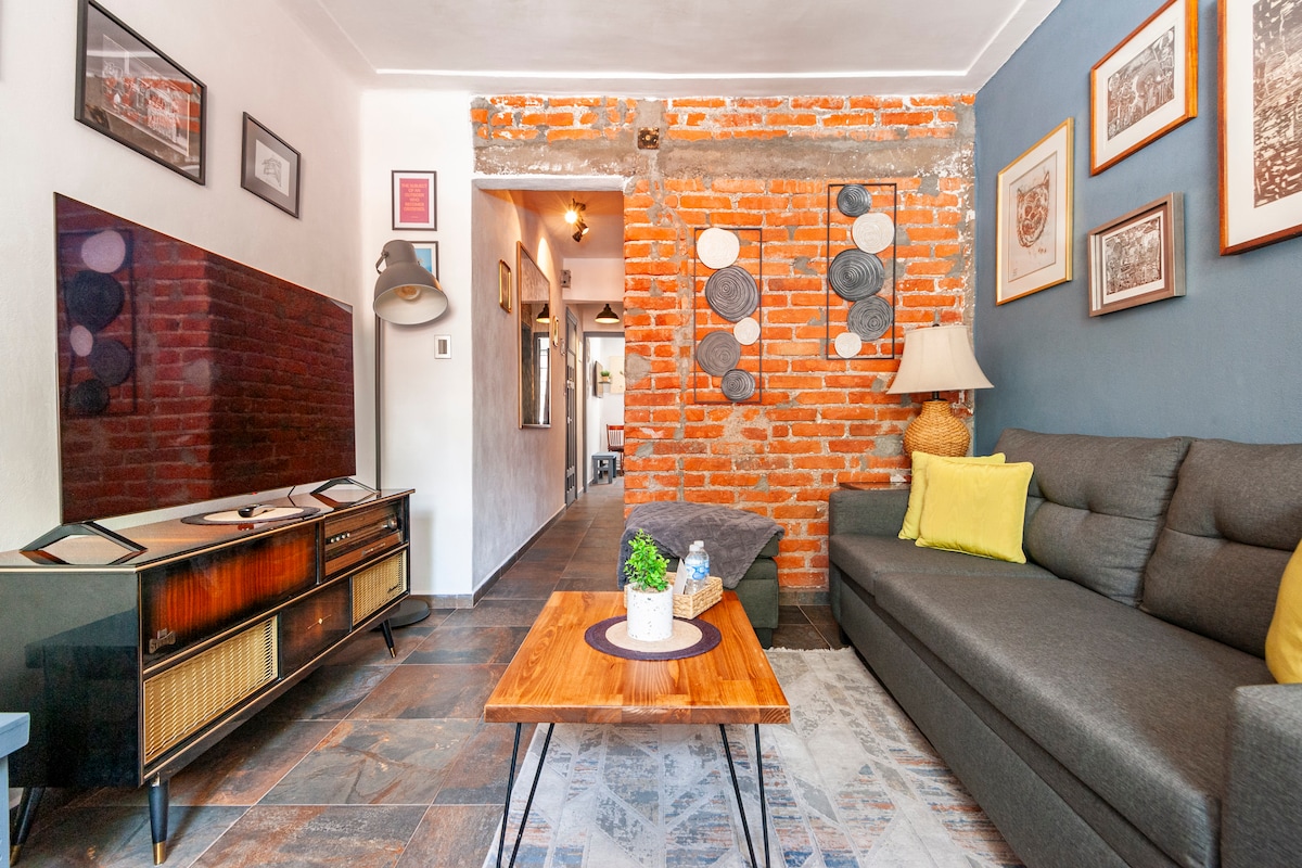 A cozy living area features a gray sofa paired with a wooden coffee table. A vintage television stands against a wall with exposed brick, while framed artwork adorns the adjacent walls. Light is provided by a stylish floor lamp, enhancing the welcoming ambiance.