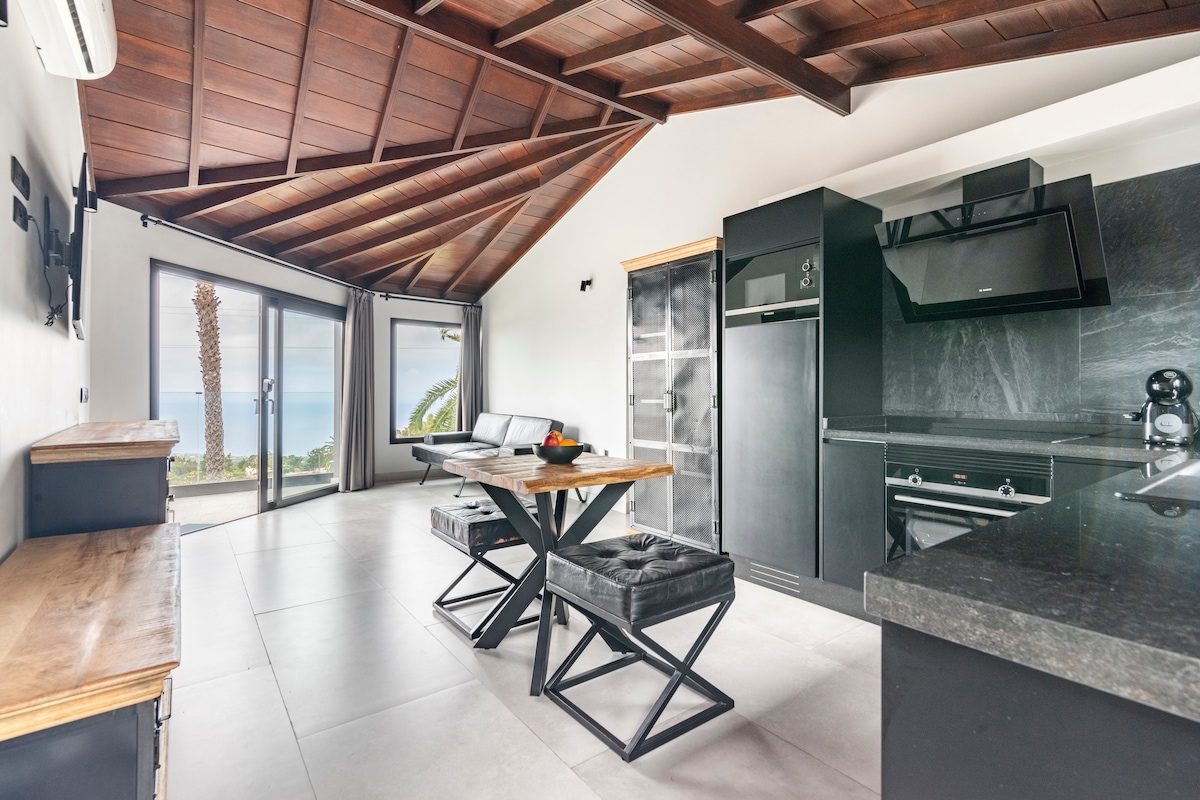 An open living and kitchen area features modern black cabinetry and a dining table with cross-legged benches. Large glass doors provide access to a terrace with sea views, complemented by wooden beams on the ceiling and a sleek, minimalist design.