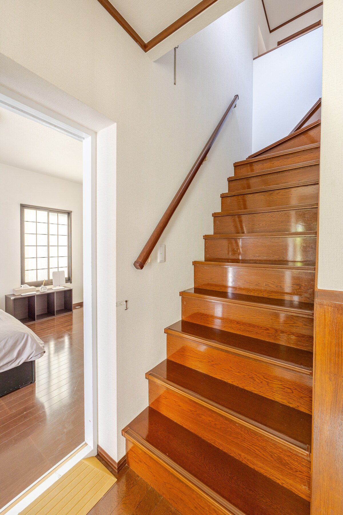 A wooden staircase leads upstairs, illuminated by natural light filtering through a nearby window. The polished steps offer a warm, inviting appearance. A doorway opens into a room visible in the background, contributing to an open and airy feel.