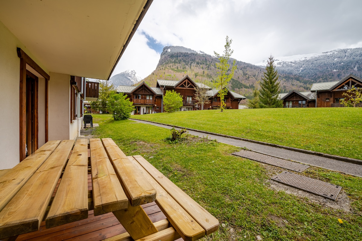 A wooden outdoor table is positioned on the patio, overlooking a grassy area with well-maintained trees and distant mountain views. The residence features a charming alpine architecture, with multiple buildings visible in the background, surrounded by nature.