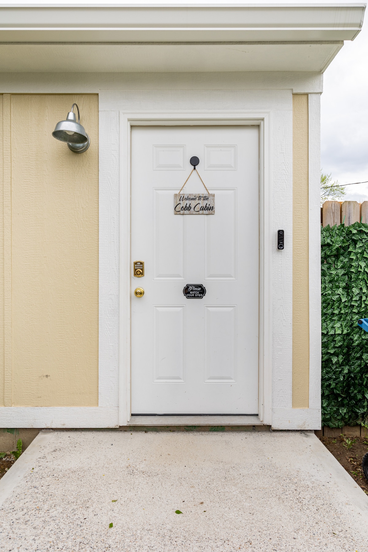 A white entry door is framed by light beige siding. A welcome sign hangs above the door handle, and a small security panel is visible beside the door. A concrete path leads to the entrance, surrounded by greenery on one side.