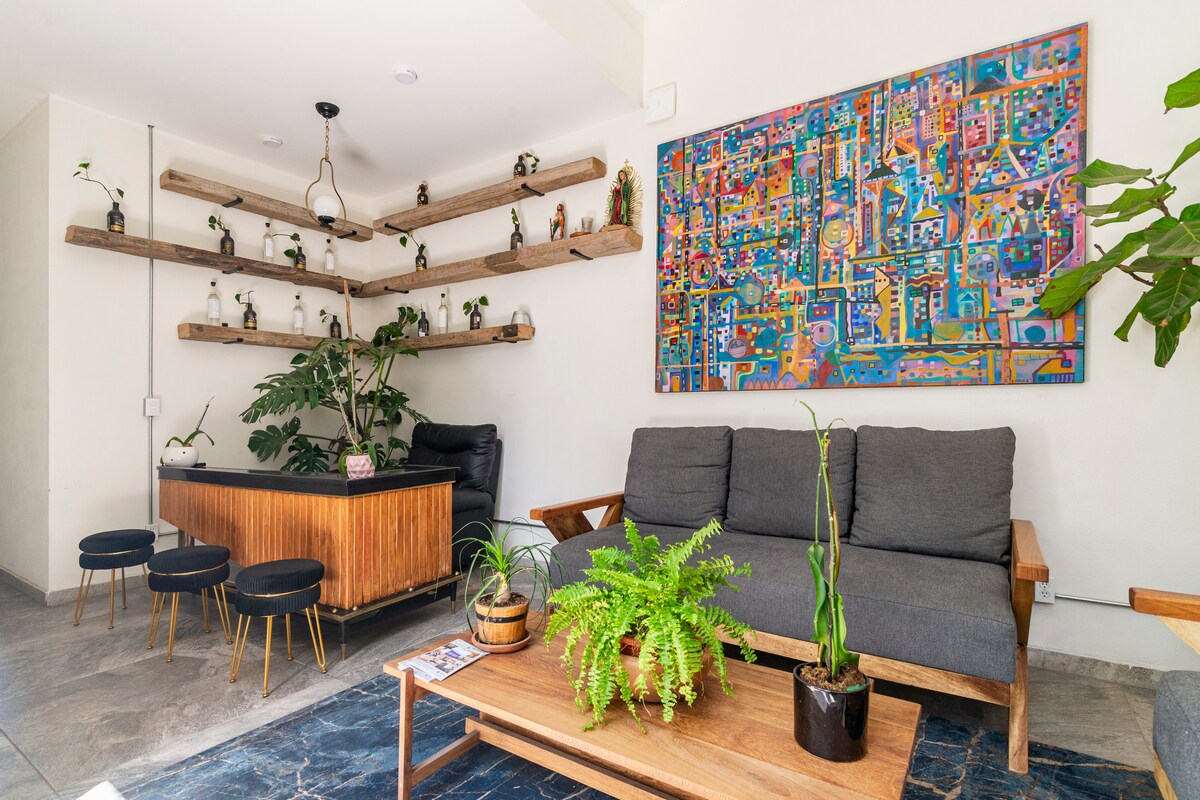 A welcoming living area is showcased, featuring a gray sofa and a wooden table. Green plants are arranged around the space, accompanied by decorative shelves with small items. A colorful artwork adorns the wall, adding a touch of vibrancy to the neutral color scheme.