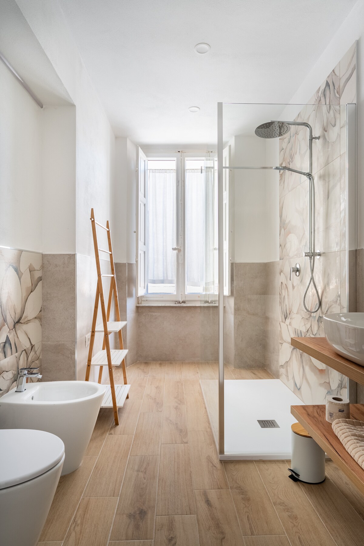 A clean and modern bathroom is displayed, featuring a spacious walk-in shower with a glass partition. A wooden ladder is positioned against the wall. Natural light enters through two windows with light, flowing curtains. A sink is positioned on a wooden shelf, complementing the neutral tones.