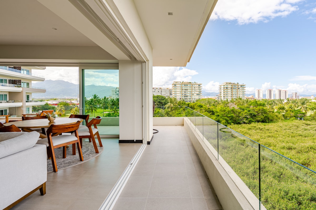 A spacious balcony offers a panoramic view of lush greenery and distant buildings. The area is furnished with a dining table and chairs, complemented by large glass doors that extend the open-concept living space. Natural light fills the interior, enhancing the welcoming environment.