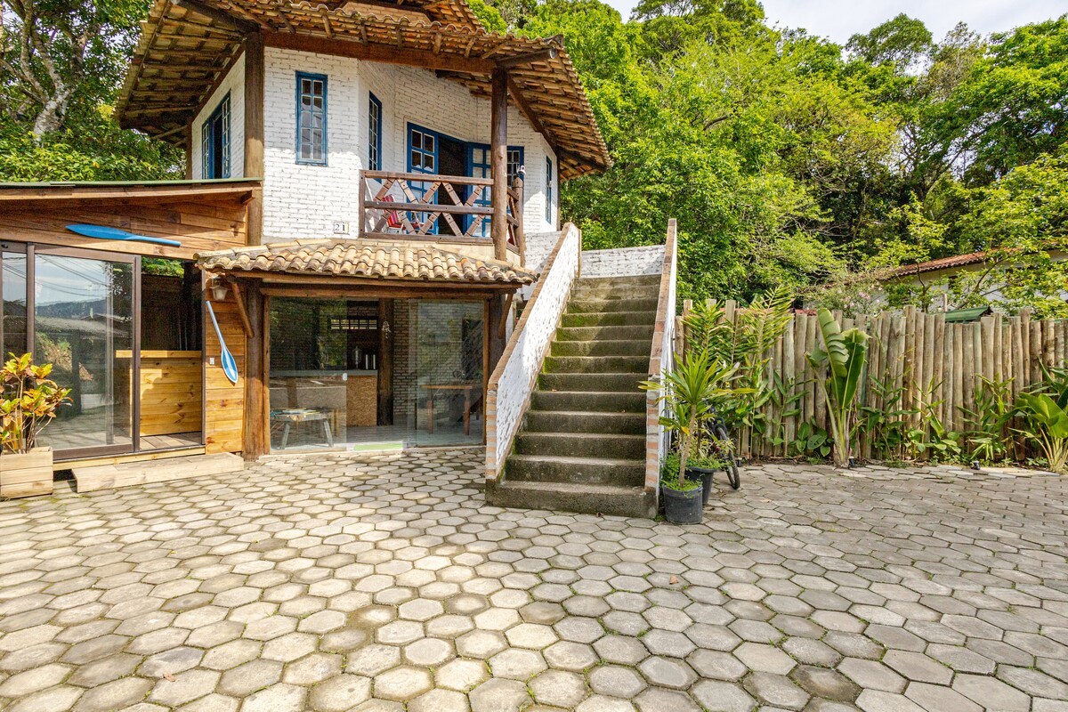 A two-story structure is framed by lush greenery, featuring a distinctive thatched roof and blue-framed windows. A broad staircase leads to the upper level, while a paved area is visible, bordered by planters and tropical plants, creating a spacious outdoor environment.