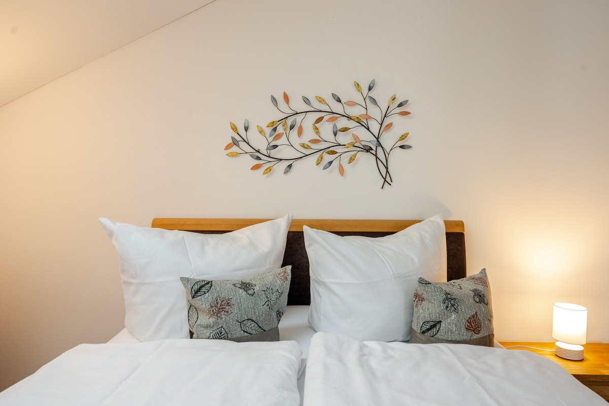 A cozy sleeping area features a spacious bed adorned with soft white linens and decorative pillows. A wall decoration of intertwined leaves adds a natural touch, while a bedside lamp provides gentle illumination. The uncluttered design creates a peaceful ambiance.
