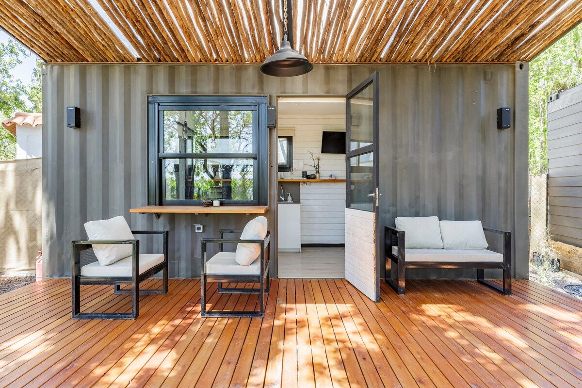 An outdoor seating area features two black armchairs and a two-seat bench, all arranged on a wooden deck. The modern structure showcases large windows and a door leading to the interior space, complemented by a decorative overhead cover made of natural materials.