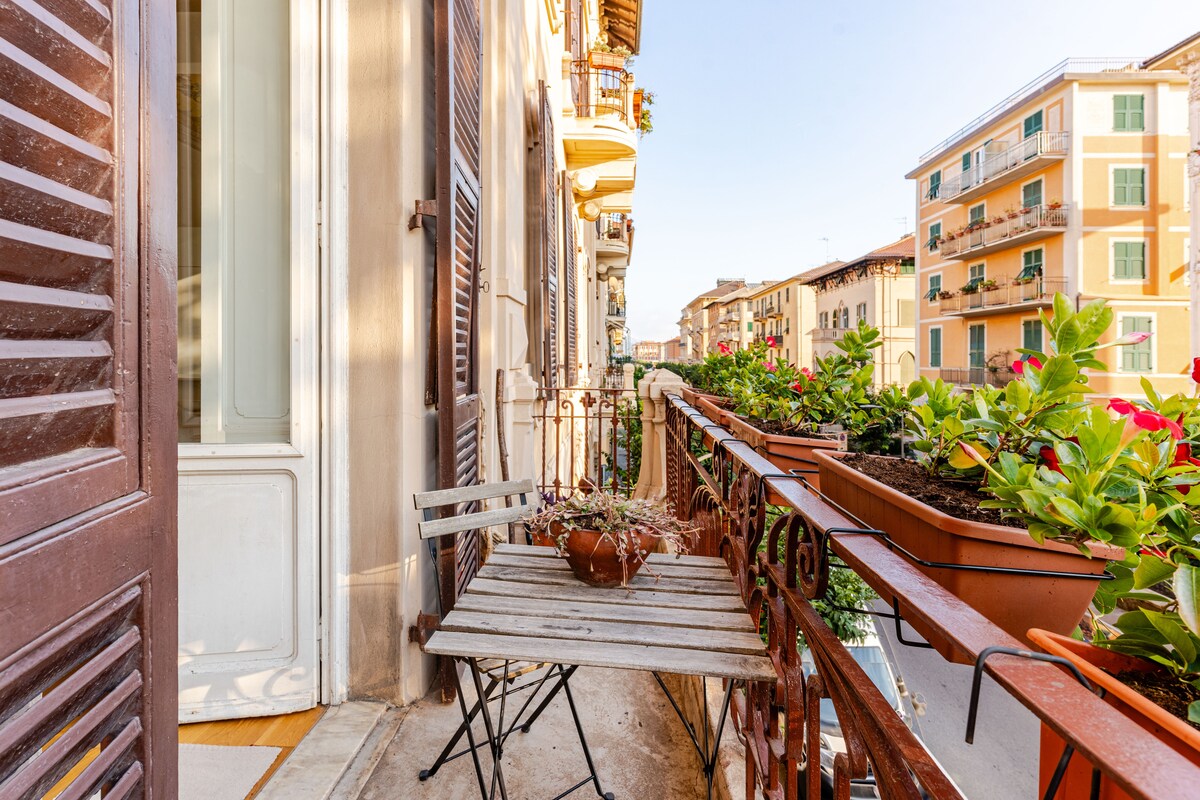 A small balcony features a wooden table with two folding chairs, surrounded by vibrant flowering plants in window boxes. Sunlight casts gentle shadows on the balcony floor, highlighting the charming details of the wrought-iron railing and the building's facade.