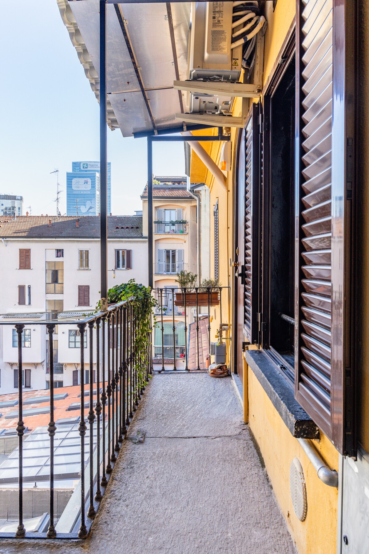 A narrow balcony is visible, featuring a wrought iron railing. Potted plants add a touch of greenery. The view includes neighboring buildings and a skyline with urban architecture, while a clear blue sky is seen overhead.